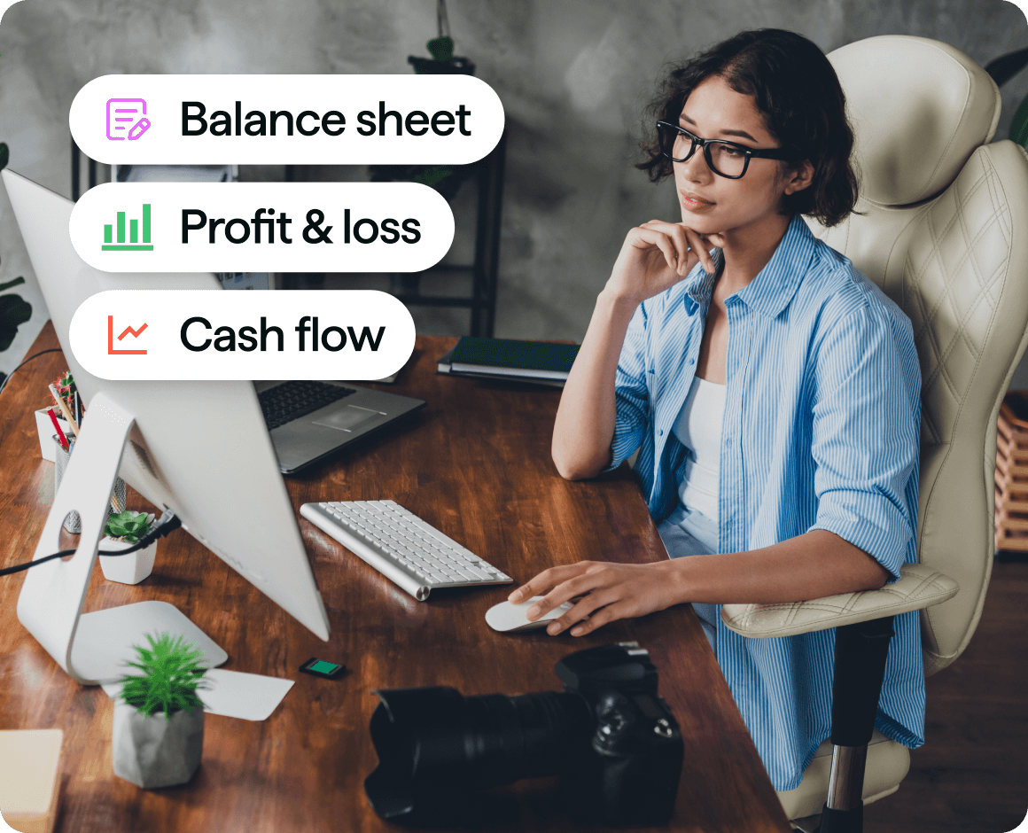 A person wearing glasses sits at a desk using a desktop computer, resting their chin on one hand while holding a mouse with the other. Overlaid labels on the image read ‘Balance sheet,’ ‘Profit & loss,’ and ‘Cash flow,’ each with an icon, alongside office items such as a keyboard, notebook, and camera on the desk.